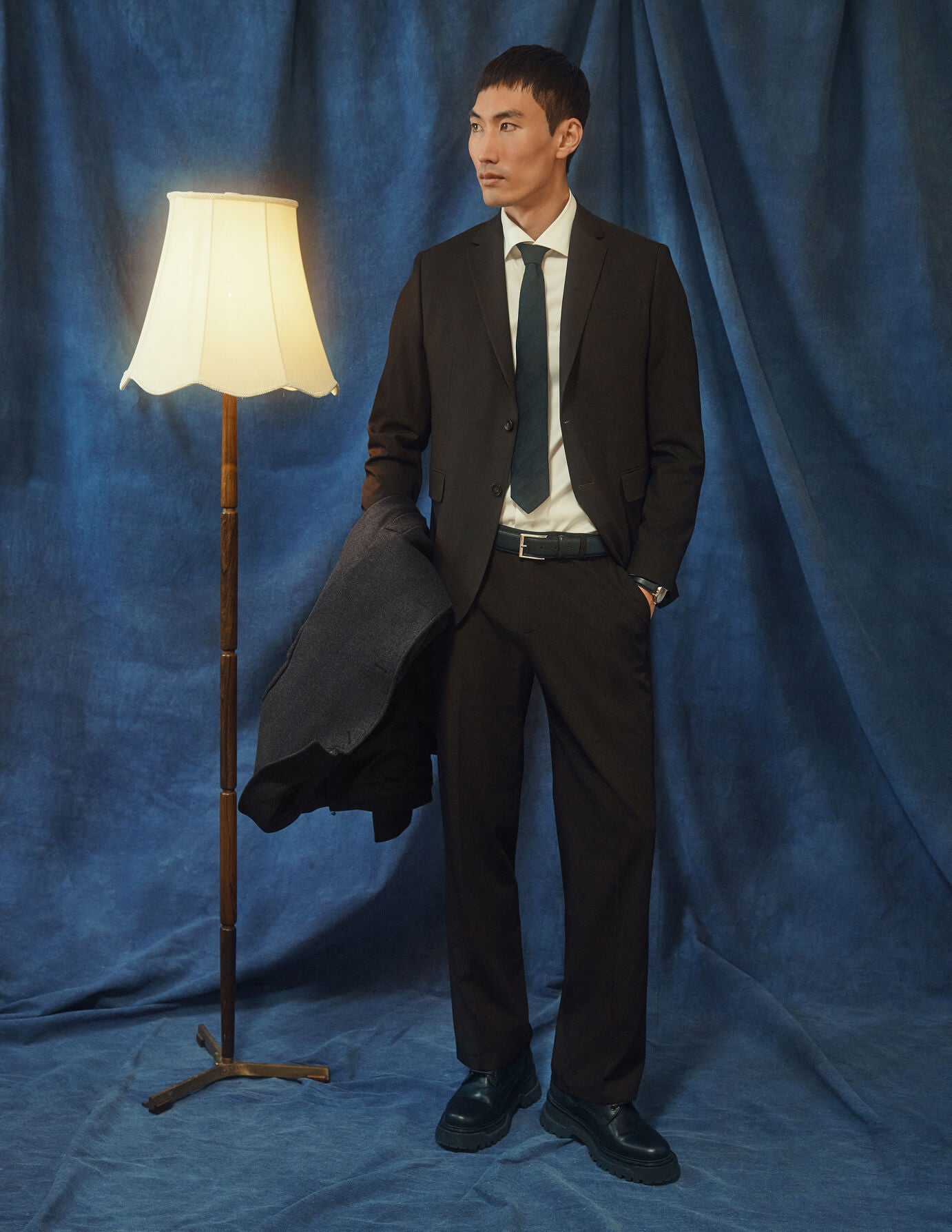Ein Mann in einem Motion Suit Pants Relaxed Cognac-Anzug, dazu ein weißes Hemd und eine Krawatte, steht neben einer Stehlampe.